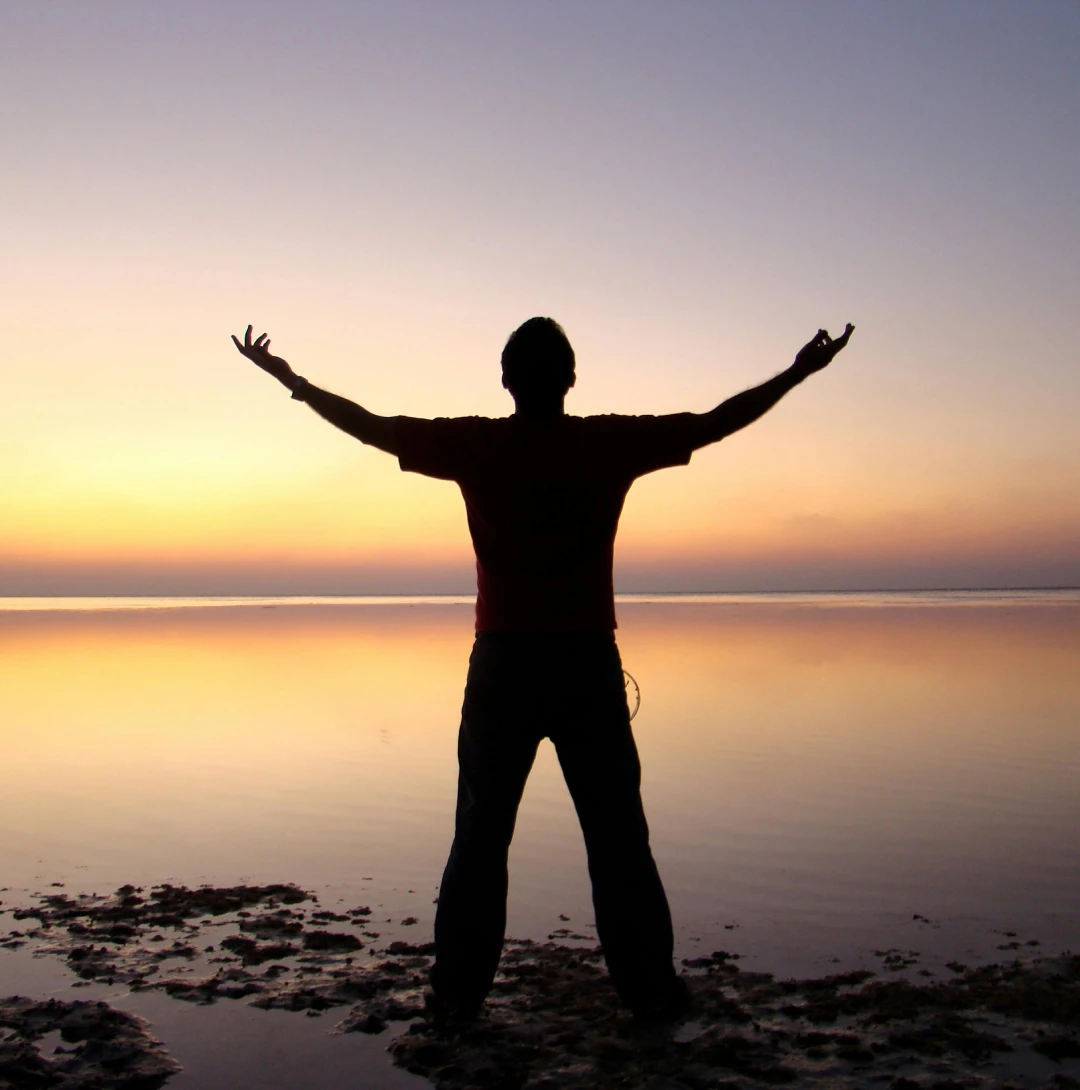 Silhouette of a person standing with arms outstretched on a beach at sunset, facing the horizon.