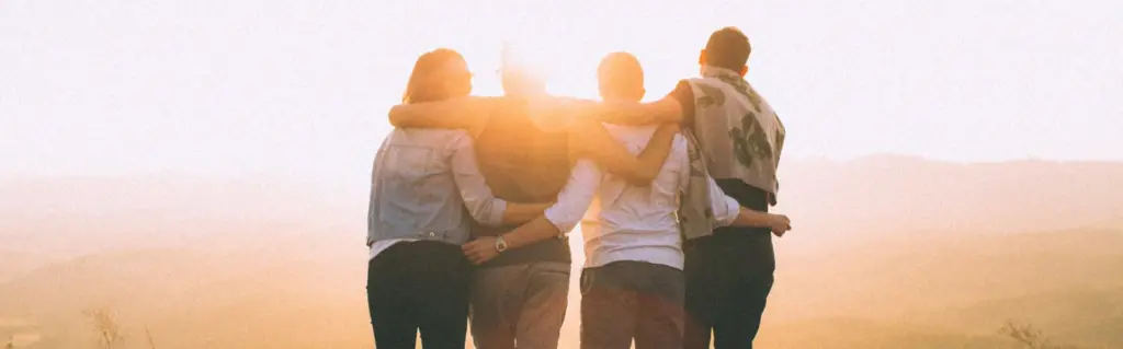 Four people stand together with their backs to the camera, arms around each other, facing a sunlit landscape.