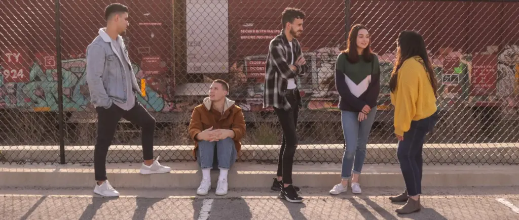 A group of five people is gathered outdoors near a chain-link fence with graffiti-covered train cars in the background. One person sits on the ground, while the others stand around chatting.