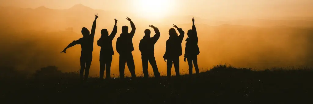 Silhouettes of six people standing on a hill at sunset, each raising one or both arms in various poses against a golden sky.