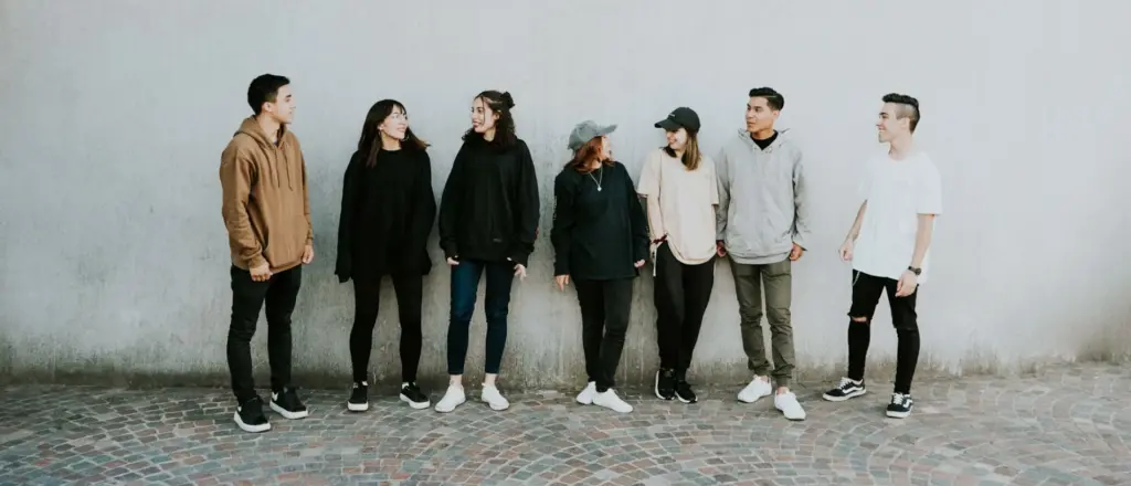 A group of seven people, casually dressed, stand in a line against a plain wall, some looking at each other and some looking forward.